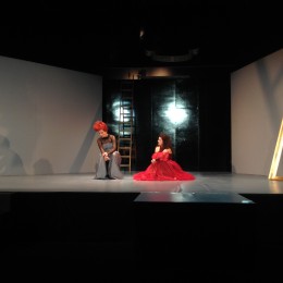 Eine Bühne auf der sich zwei Frauen befinden. Beide auf dem Boden kniend. Die eine trägt ein rotes Kleid, die andere hat im Gegenzug rote Haare.