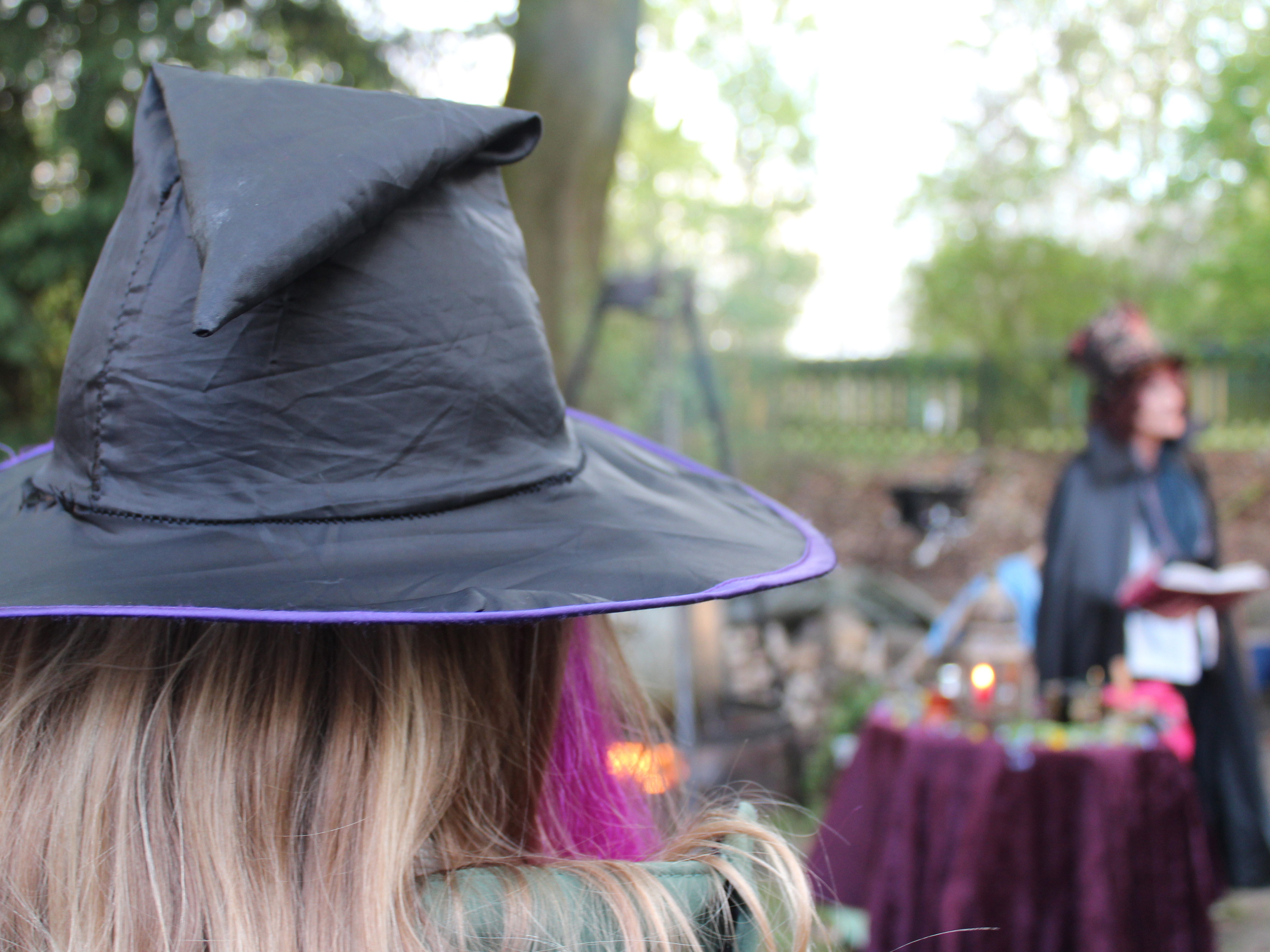 Es ist ein Kind mit einem Hexenhut von hinten zu sehen. Im Hintergrund befindet sich verschwommen eine Hexe die aus ihrem Buch liest.