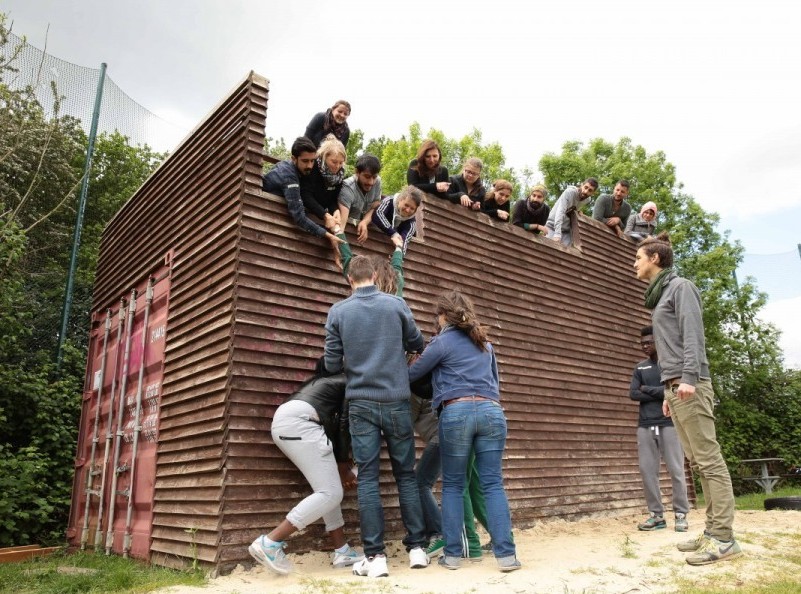 Zu sehen ist eine Gruppe die vor einer hölzernen Mauer steht. Einige befinden sich über der Mauer.