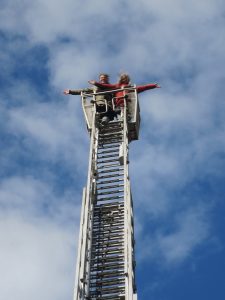Zwei Frauen am oberen Ende einer Feuerwehrleiter vor blauem Himmel