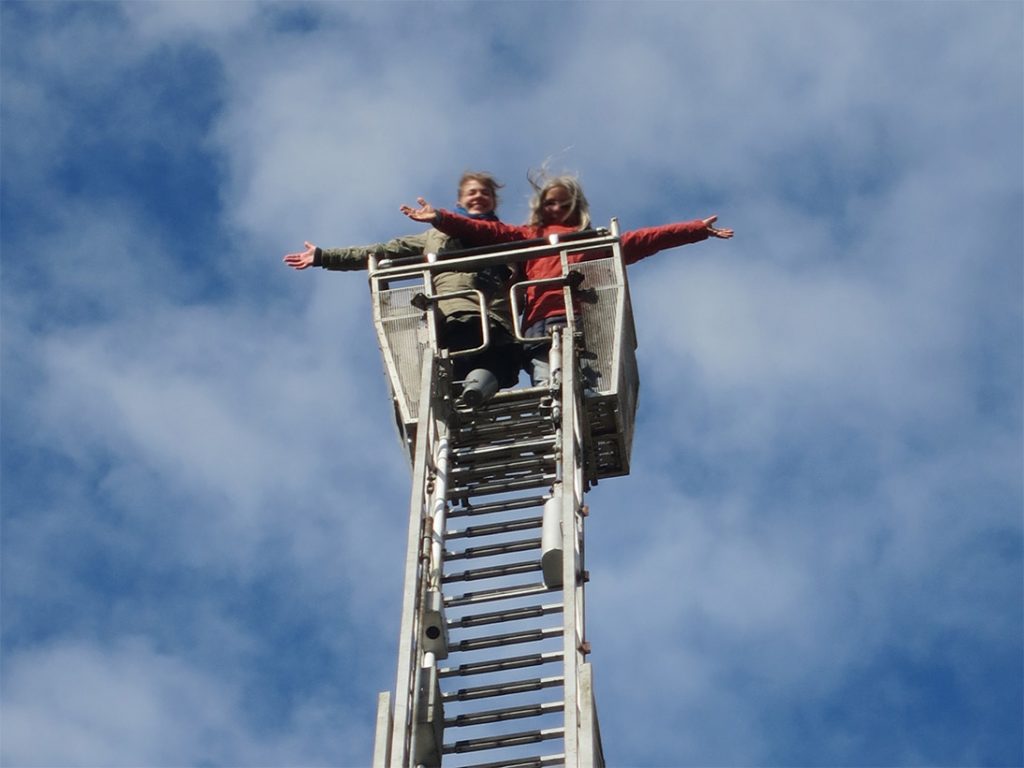Zwei Frauen am oberen Ende einer Feuerwehrleiter vor blauem Himmel