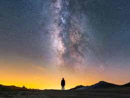 Person steht in der Wüste, über ihr der Sternenhimmel und die Milchstraße. Der Himmel leuchtet gelb und die zentral stehende Person ist nur als Silhouette zu erkennen.