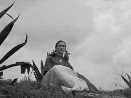 Frida Kahlo sitzt in der Natur auf dem Boden, schaut in die Kamera. Schwarz weißes Foto.