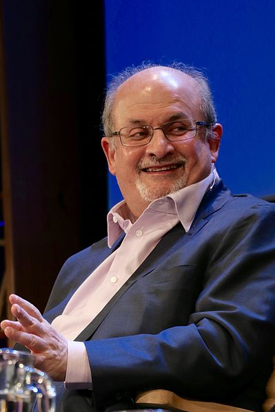 Der Autor Salman Rushdie sitzt auf einem Stuhl vor einem blauien Hintergrund während eines Interviews. Er hat einen grauen Haarkranz, eine Halbglatze, ein schwarzes Jacket, weißes Hemd und eine Brille.