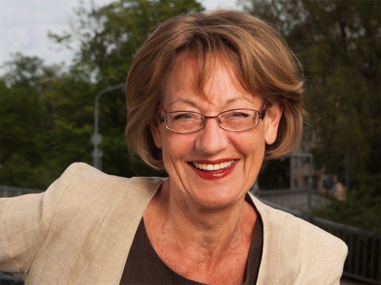 Portrait einer Politikerin mit Brille und rot-geschminkten Lippen. Sie hat braune Haare und trägt ein beiges Jacket. Gudrun Schyman.