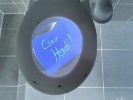 Eine Toilette von oben fotographiert. Sie ist geöffnet. Ein blaues Papier mit der Aufschrift ' Ciao Heidi' liegt darin