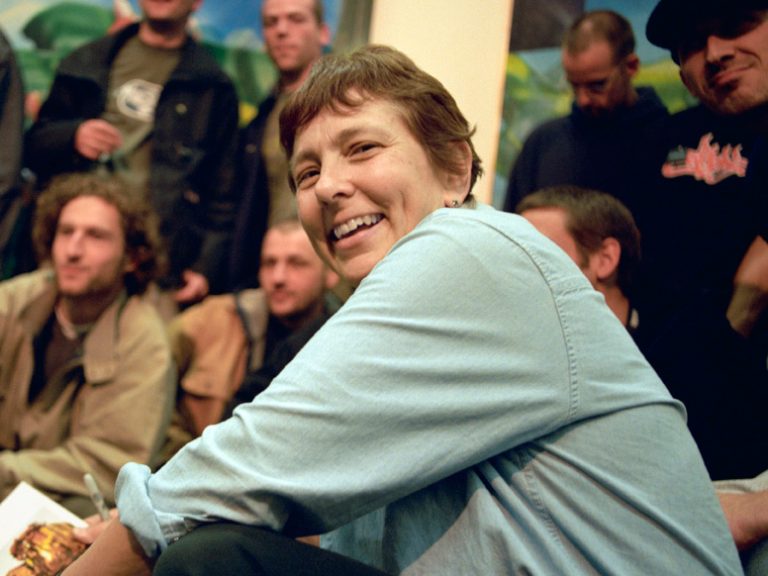 Martha Cooper, lächelnde Frau sitzend im Vordergrund, hinter ihr unscharfe Menschen