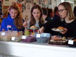 feministisches Frühstück, Drei frauen nehmen sich Speisen bei einem Frühstücksbuffet. Im Hintergrund sind weitere Frauen, die sich Unterhalten. Eine Wand im Hintergrund ist mit Plakaten behangen.