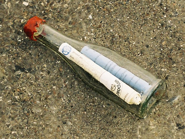 Glasflasche auf Sanboden liegend mit zwei beschrifteten Papierrollen im inneren der Flasche