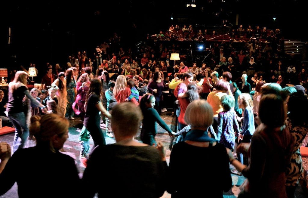 Eine Gruppe tanzender Frauen im Hintergrund ein Orchester beim Fest der Frauen*