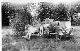 Schwarz-Weiß Bild von einer Frau, einem Mann und einem Kind, die auf Liegestühlen in einem Garten liegen. Der Mann liest eine Zeitung.