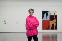 Ulrike Müller steht in einem pinken Pullover vor ihren Kunstwerken die an einer Wand hängen
