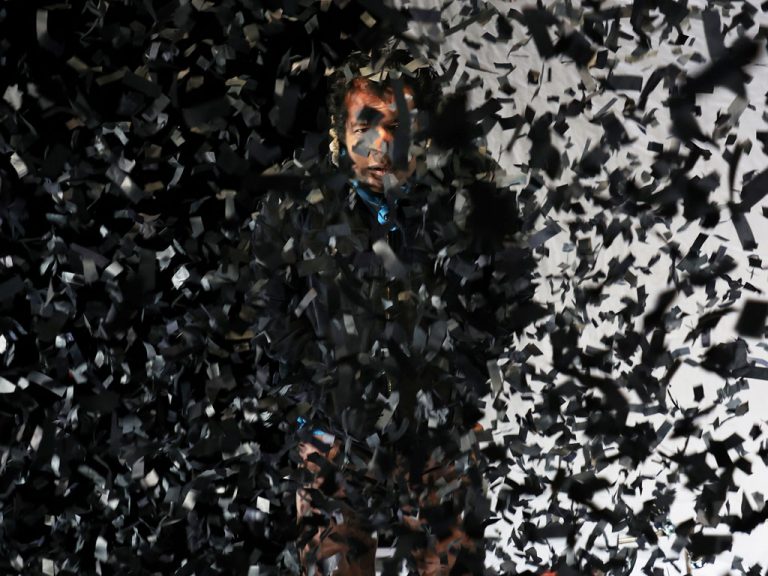 Patrick Balaraj Yogarajan als Wahab in "Im Herzen tickt eine Bombe" von Wajdi Mouawad auf der Bühne am Theater Bremen steht hinter aufgewirbelten schwarzen Papierschnipseln