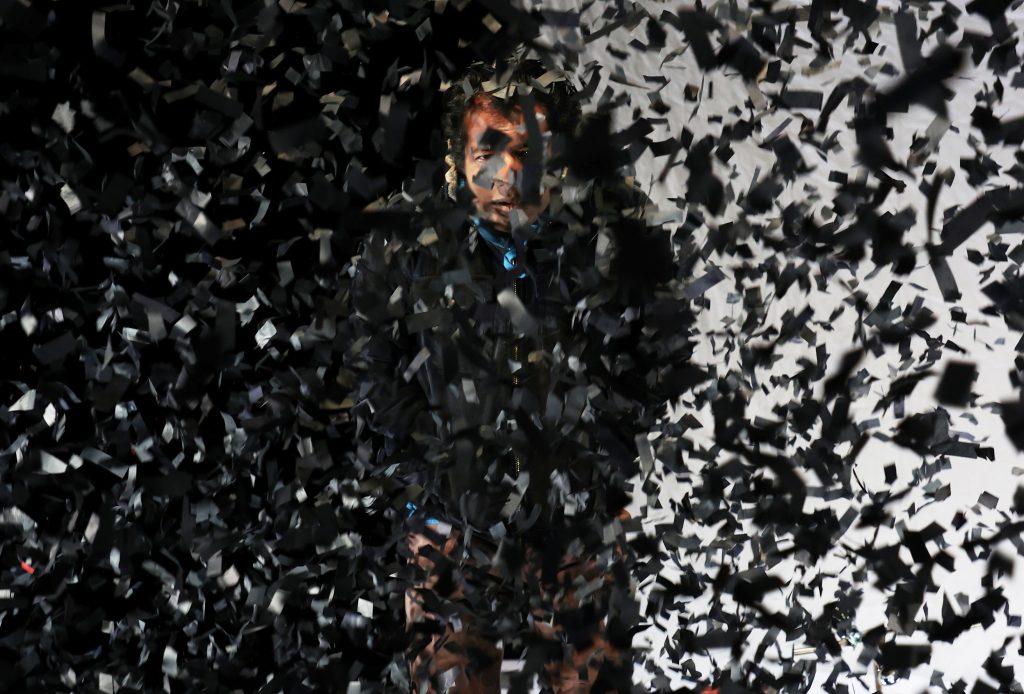 Patrick Balaraj Yogarajan als Wahab in "Im Herzen tickt eine Bombe" von Wajdi Mouawad auf der Bühne am Theater Bremen steht hinter aufgewirbelten schwarzen Papierschnipseln
