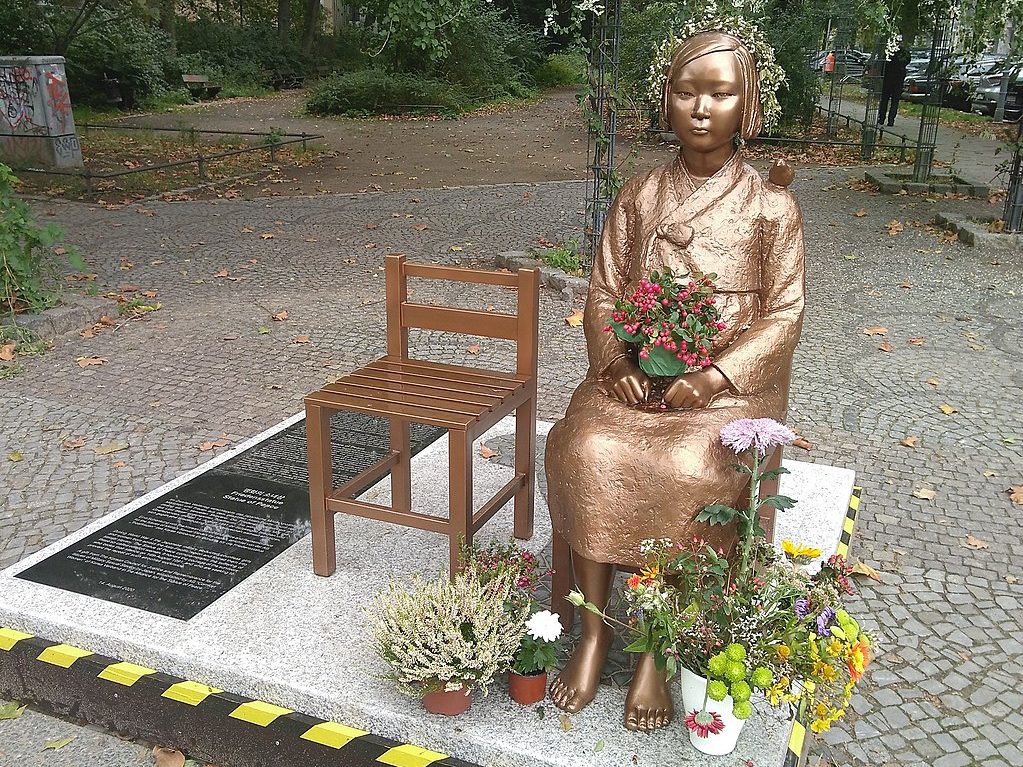 Die Bronzestatue eines Mädchens sitzt auf einem Holzstuhl auf einer Plattform aus weißem Marmor. Sie trägt traditionell koreanische Kleidung und hat echte Blumen im Schoß und zu ihren Füßen drapiert