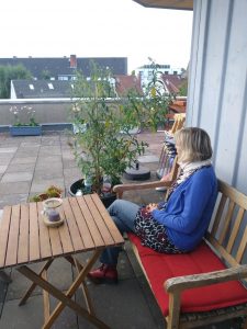 Eine Frau mit blauer Strickjacke sitzt auf einer Dachterrasse auf einer Bank. Sie blickt in die Ferne, neben ihr ist ein kleiner Holztisch und einige Topfpflanzen