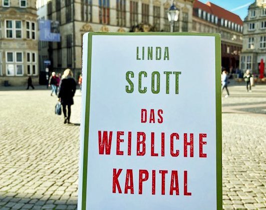 Das Buch "Das weibliche Kapital" wird hochgehalten. Im Hintergrund ist der Bremer Marktplatz. Eine Frau geht von der Kamera weg, weitere Menschen sind im Hintergrund zu erkennen