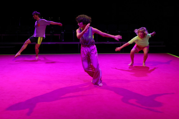 Drei Personen bewegen sich tanzend auf einem pinken Boden. Ihre Schatten sind dabei deutlich zu erkennen