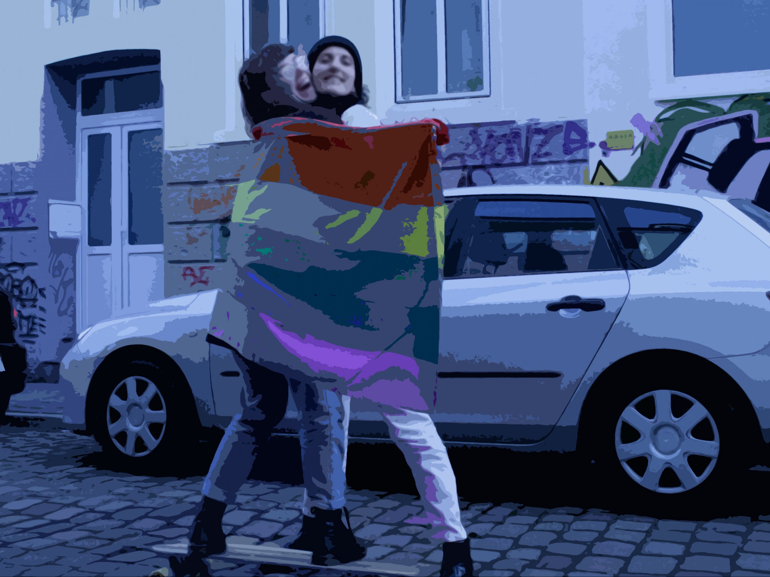 Auf der Straße. Zwei Personen umarmen sich, die hintere, größere hält eine GayPride-Flagge um beide. Die vordere, kleinere steht auf einem Longboard, sodass beide gleich groß sind. Beide lachen.