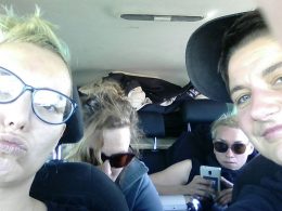 Foto von vier Personen im Auto. Zwei Personen sitzen vorne, schauen direkt in die Kamera. Zwei weitere Personen sitzen hinten und tragen eine Sonnenbrille. Eine schaut nach unten, die andere auf ihr Handy.