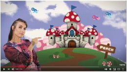 Sukini zeigt auf eine Burg, die Fliegenpilze als Türme hat, die Burg steht auf einem pixeligen Hügel und der Hintergrund besteht aus pixeligen Wolken. Ein Schild, auf dem Peach steht, zeigt zur Burg