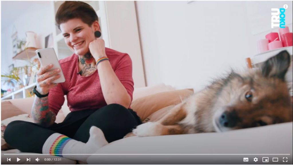 Eine Frau sitzt links im Foto im Schneidersitz auf einem Sofa und guckt lachend auf ihr Handy. Neben ihr recht im Bild liegt ein kleiner Hund mit braunem Fell auf dem Sofa und blickt den*der Betrachter*in des Fotos ins Auge.