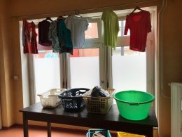 Wäsche hängt auf Bügeln vor einem Fenster, auf einem Tischen stehen Wäschekörbe und Kleidung