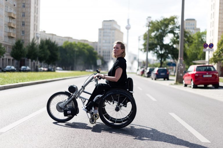 Junge Frau im dreirädrigen Rollstuhl, der wie ein Motorrad aussieht auf einer großen Straße in Berlin mit dem Fernsehturm im Hintergrund