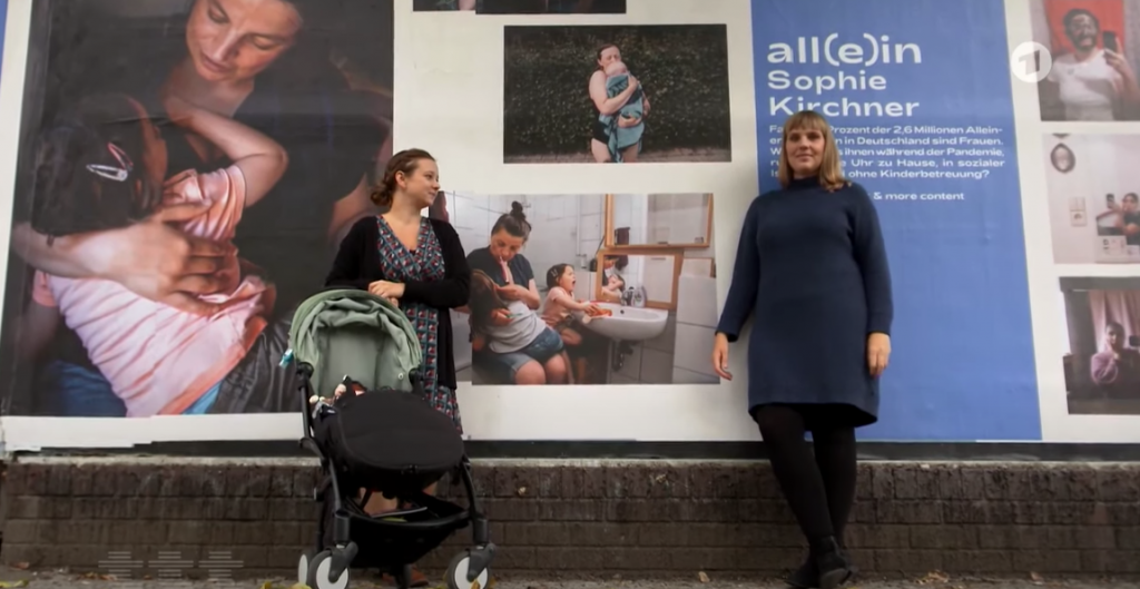 Zwei Künstlerinnen stehen vor einer Plakatwand, wo ihre Fotografien über alleinerziehende Mütter abgebildet sind.