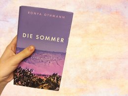 Das Buch "Die Sommer" von Ronya Othmann wird von einer Hand gehalten. Das Buch ist lila und ziegt eine syrische Berglandschaft. Der Hintergrund ist geldb/pink miliert.