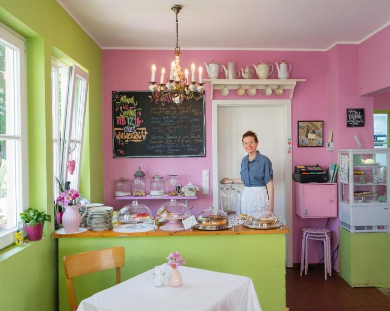 Café Radieschen, Frau hinter der Kuchentheke, rosa und grüne Wände, Tisch mit Stuhl im Vordergrund, Blumenvase auf Tisch