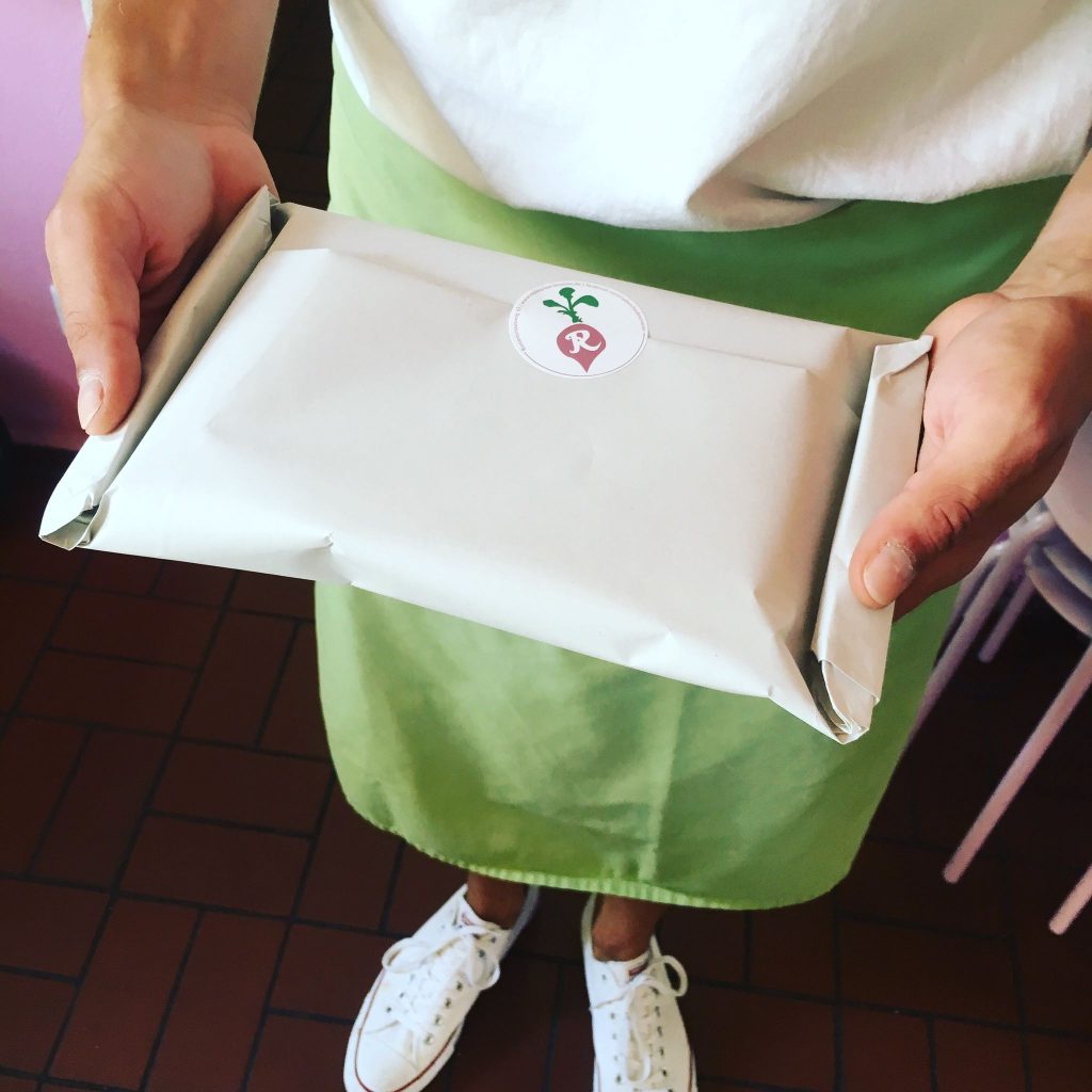 Person mit grüner Schürze, weißes Oberteil, weiße Schuhe, verpackten Kuchen in der Hand, Radieschen-Sticker auf Verpackung