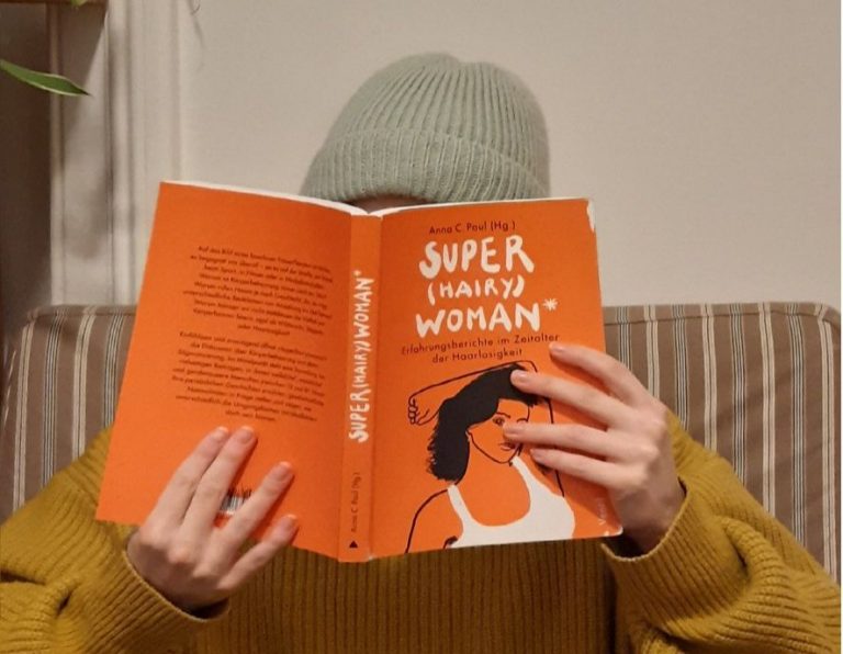 Person im gelben Pulli, hält ein oranges Buch mit der Aufschrift "Super hairy Woman"
