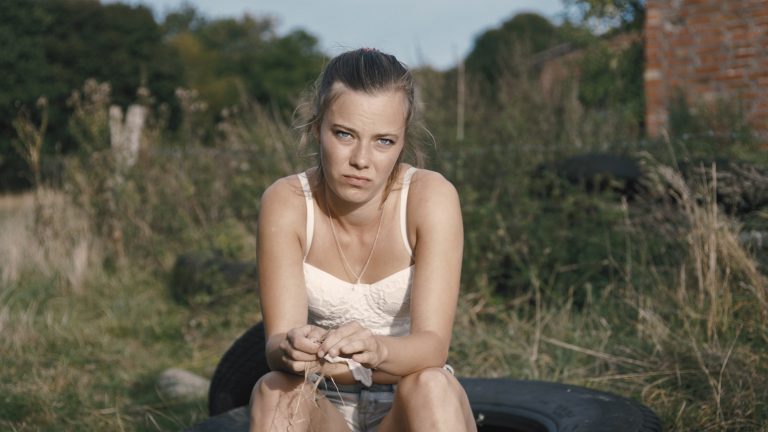 Christin, gespielt von Saskia Rosendahl sitzt auf einem großen Traktorreifen und schaut gelangweilt hinter die Kamera. Mit den Händen spielt sie mit einem Grashalm. Es ist ein heißer Sommertag, sie trägt ein weißes kurze Top.