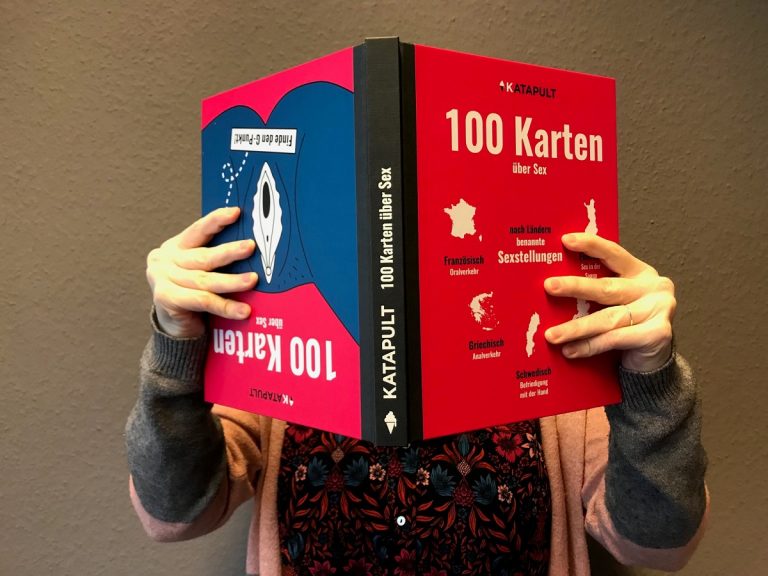 Person hält das Buch "100 Karten über Sex" aufgeschlagen vor dem Gesicht, grauer Hintergrund