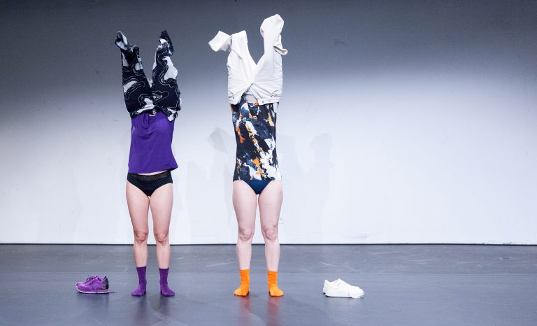 Die beiden Perfomancekünstlerinnen Tiina Sööt und Dorothea Zeyringer stehen auf der Bühne mit nackten Beinen und ihren Hosen über ihre Köpfe gestülpt als würden sie versuchen sie als Kleid überzustreifen. Das Foto ist Teil ihrer Performance Angry Hour.