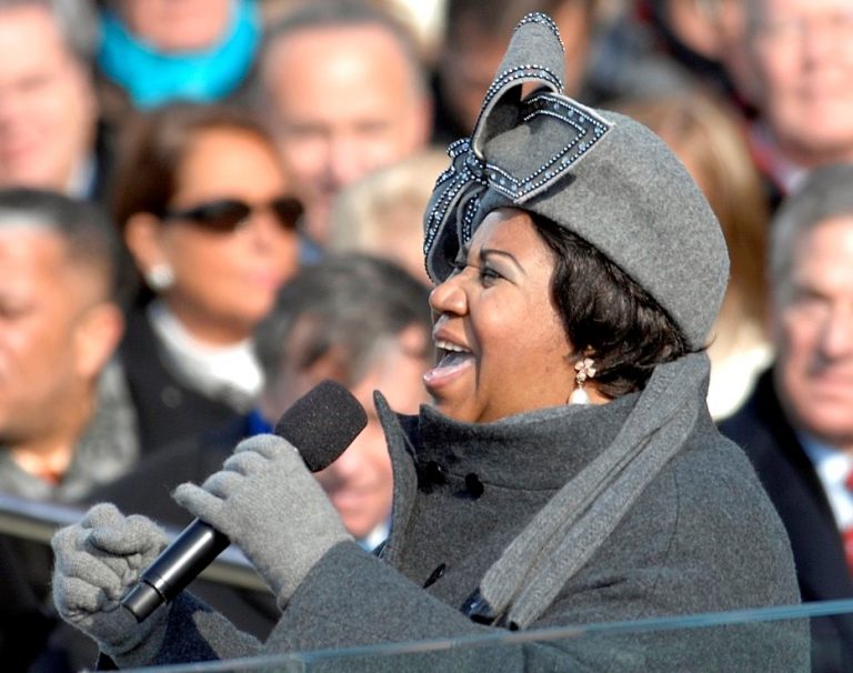 Aretha Franklin mit Mikrofon in der Hand und gekleidet in grauem Mantel, grauen Handschuhen und einem grauen Hut mit Schleife. Sie singt in ein Mikrofon, das sie in der Hand hält.