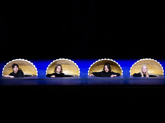 Vier Frauen in schwarzer Kleidung schauen aus golden verzierten Löchern im Boden einer Theaterbühne hervor
