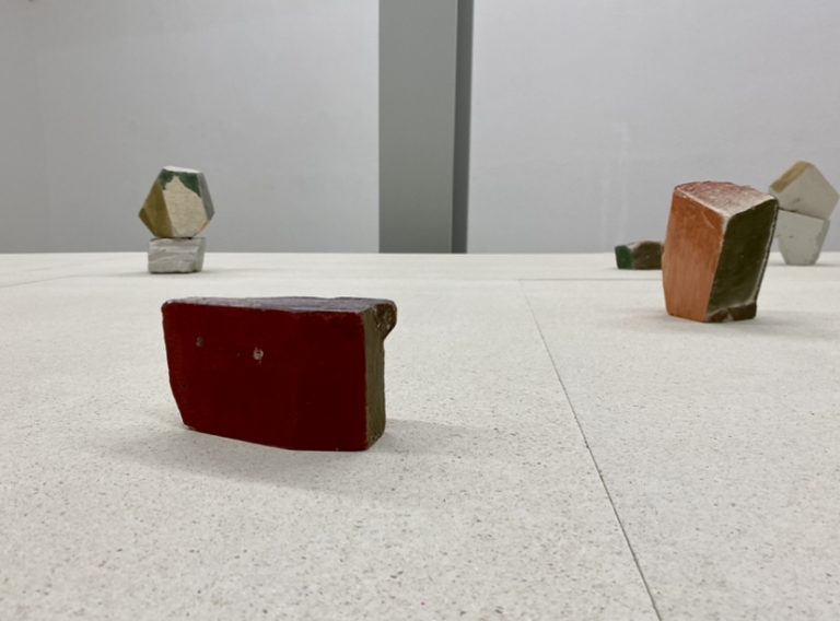 Steine, bemalt in verschiedenen, bunten Pastellfarben, sind auf einer Tischplatte verteilt ausgestellt, der Hintergrund ist schlicht.
