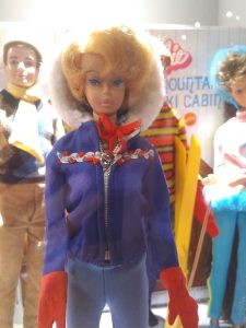 Barbie-Puppe im blauen Anorak und roten Handschuhen, im Hintergrund weitere Puppen