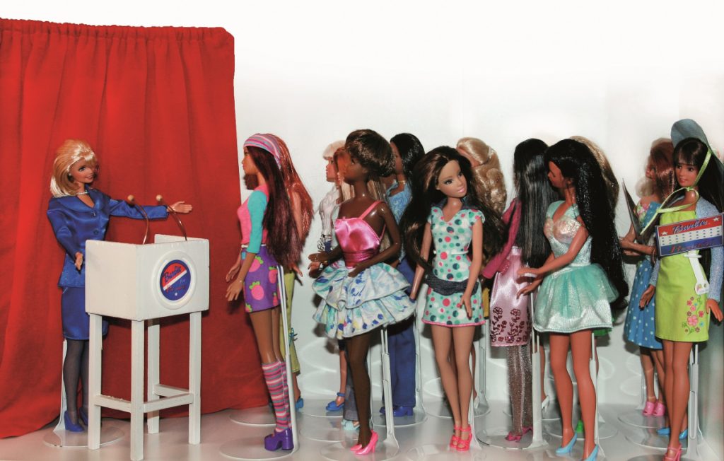 Barbie als Präsidentin am Pult vor vielen anderen Barbies