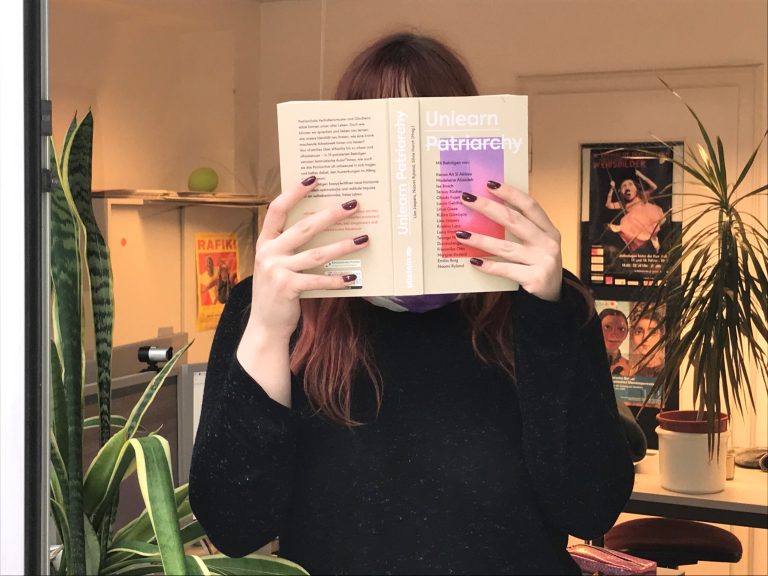 Ein Mädchen steht mit dem Buch "Unlearn Patriarchy" in einem Bürozimmer mit Pflanzen im Hintergrund