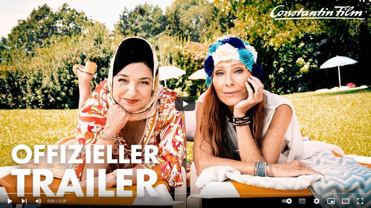 Trailerbild vom Film Freibad, auf dem zwei Frauen mit Kopfbedeckungen auf einer Decke auf einer Wiese liegen und in die Kamera grinsen