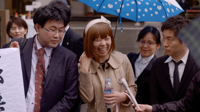 Rokudenashiko steht im Mittelpunkt des Bildes und lacht. Umgeben ist sie von Reportern und anderen Menschen