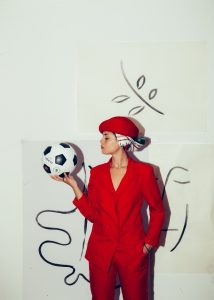 weiblich gelesene Person in rot gekleidet mit Fußball in der Hand und Wandbemalung im Hintergrund 