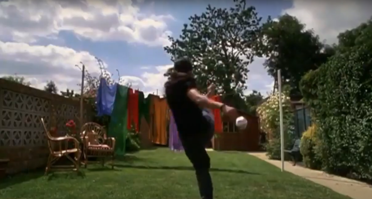 Person die einen Fußball in die Wäscheleine mit bunten Klamotten schießt. Im Hintergrund steht rechts ein Baum, der Grund ist eine grüne Wiese