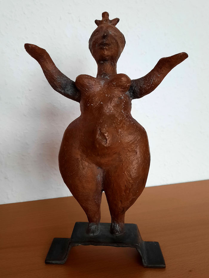 Bild einer Bronzeskulptur namens kleine Aphrodite. Die stehende Figur blickt mit erhobenen Armen nach oben.