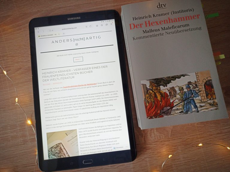 Tablet mit der Webseite von ANDERS[nicht]ARTIG. Rechts liegt eine Ausgabe des Buchs Malleus Maleficarum