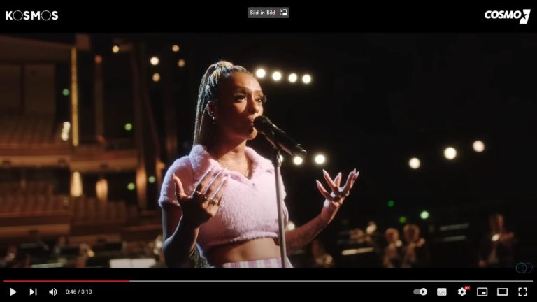 Screenshot eines Youtube Videos. Zu sehen ist die Rapperin Nura Habib Omer wie sie am Mikrofon steht und singt. Im Hintergrund ist ein Orchester zu erkennen.
