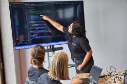 Im Rahmen eines IT-Workshops zeigt eine Frau zwei anderen Frauen auf einem großen Bildschirm Programmierbefehle